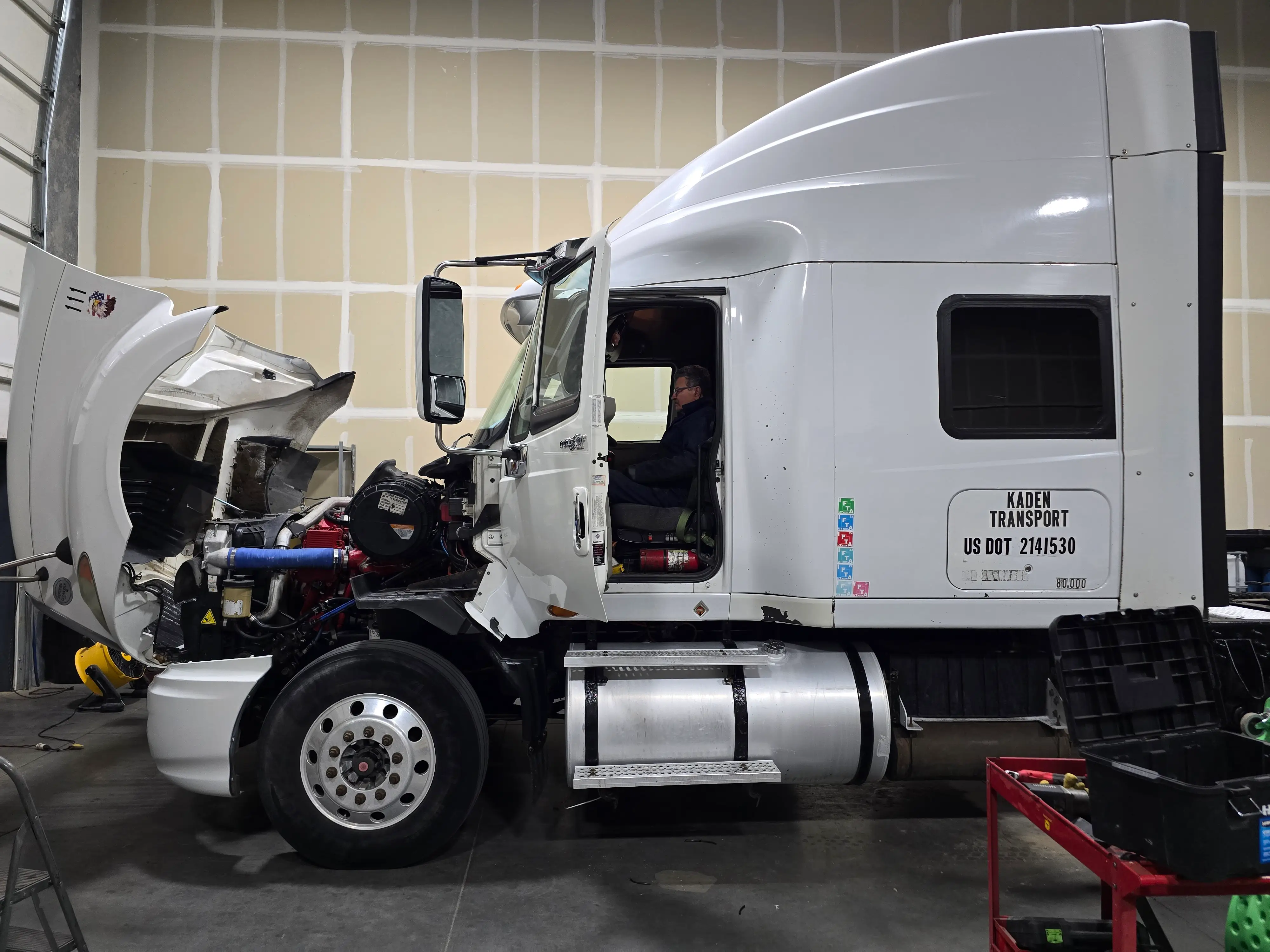 A Kaden Transport tractor in the bay with the hood tilted forward, engine exposed, and a technician sitting in the cab.