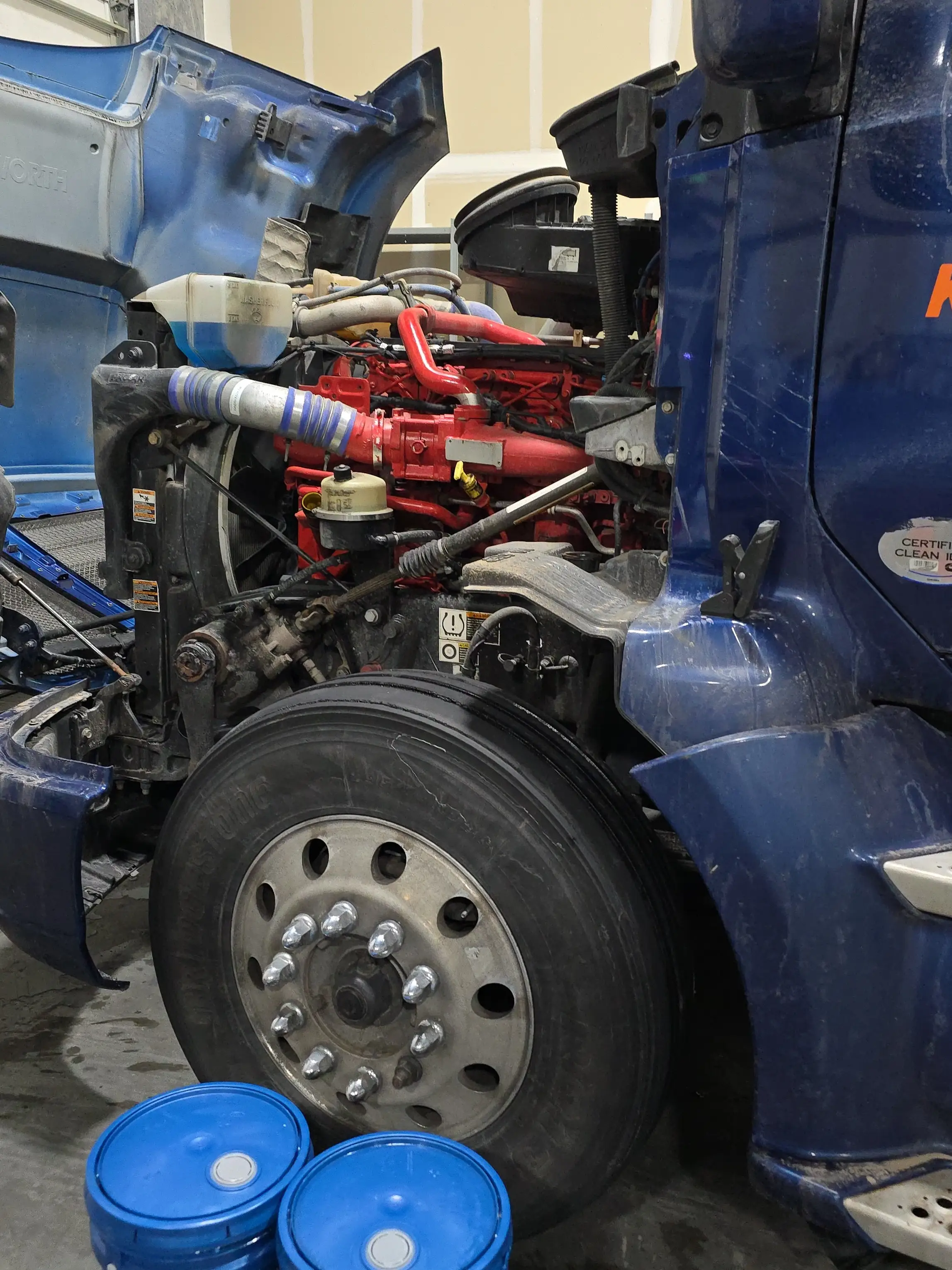 A Kenworth with the hood open in the bay, exposing a red Cummins engine, cooling hoses and the steer axle.
