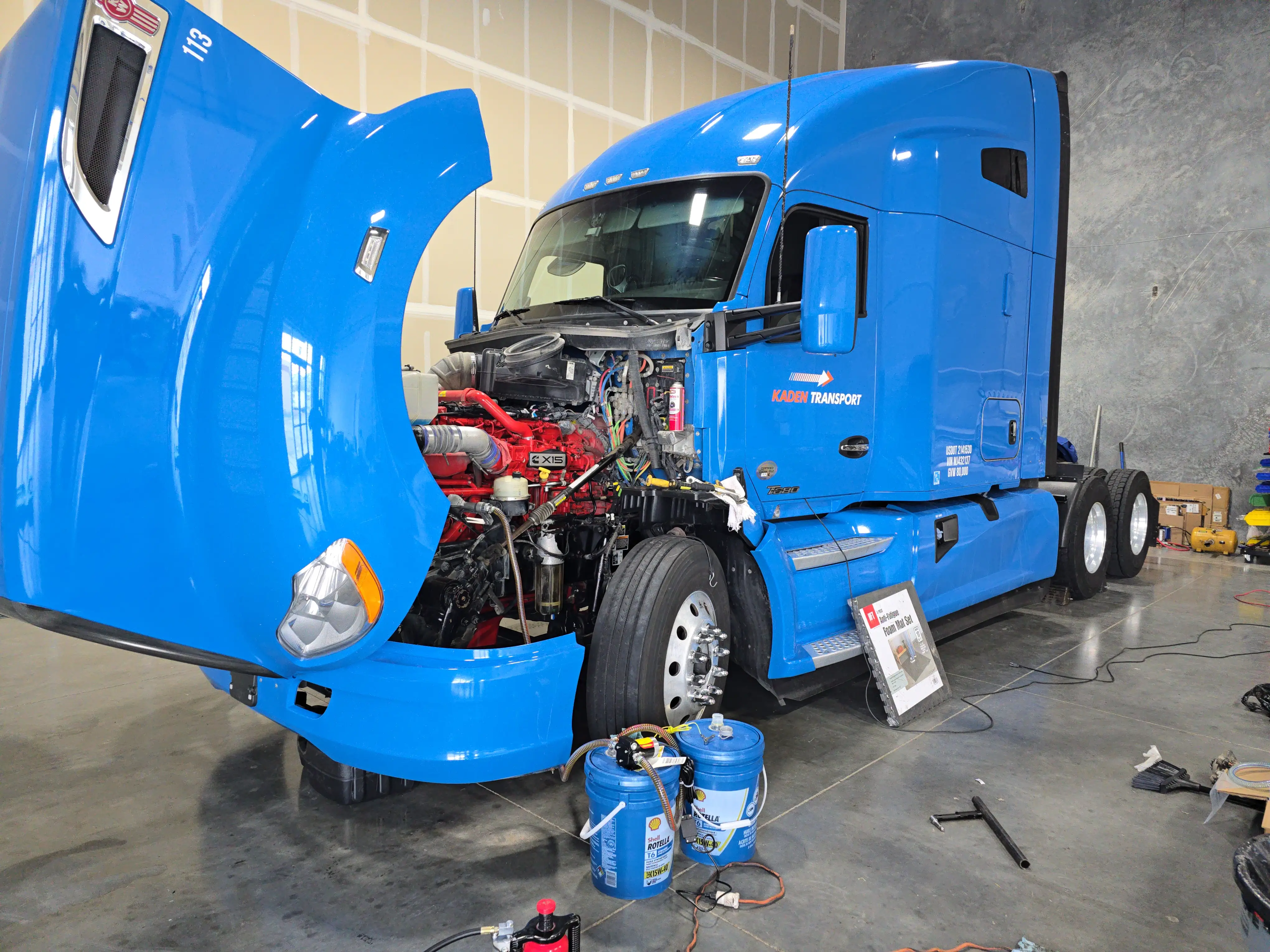 A blue Kenworth T680 in the bay with the hood tilted forward, exposing a red Cummins X15, with oil pails and tools on the floor.