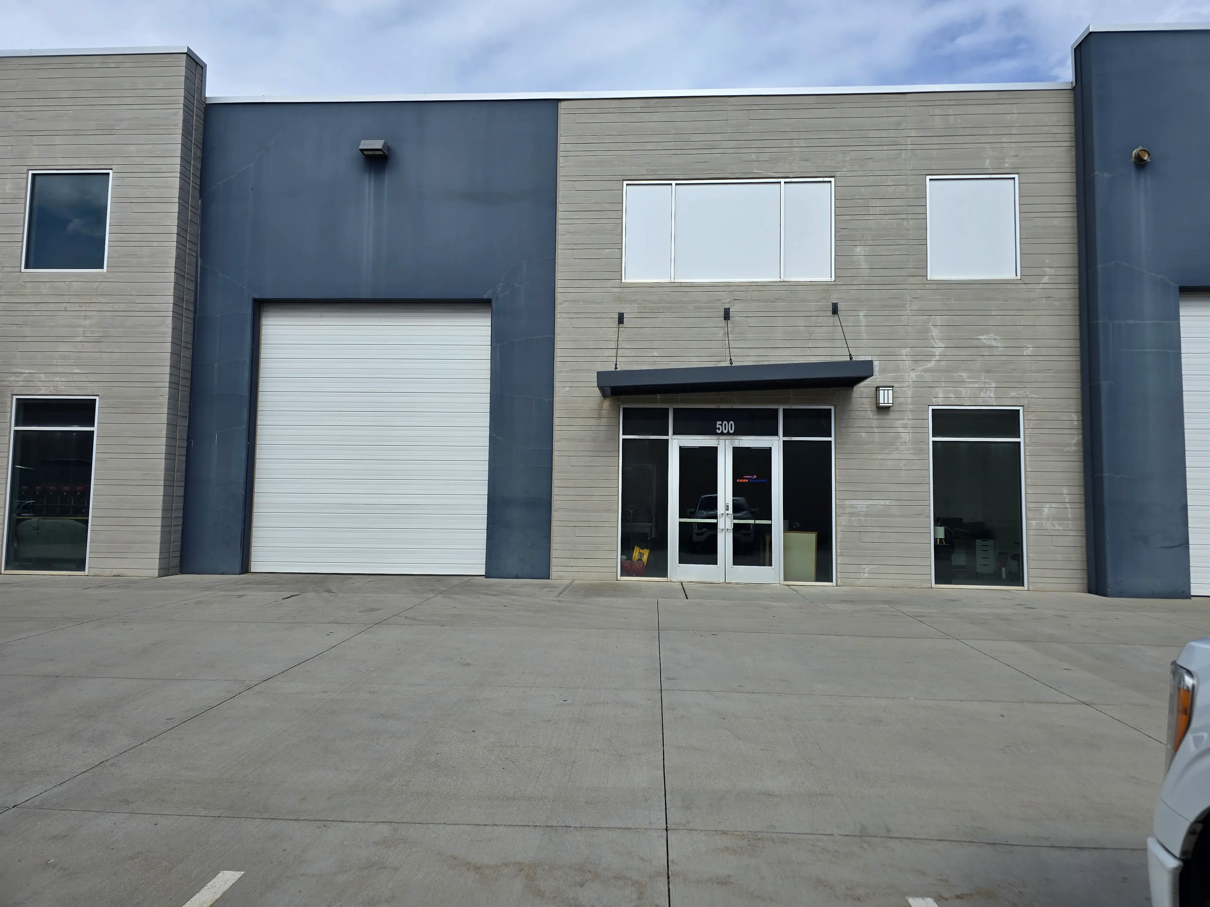 The shop from the parking area — a grey industrial unit with a white roller door to the left of the glass entrance.